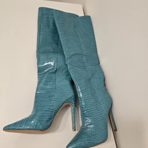 Turkosa stövlar med krokopräglat mönster - Statement-stövlar i turkost med krokodilpräglat mönster och spetsig tå. Hög klack och slank siluett, tillverkade i glansigt syntetmaterial. Perfekta för dig som vill sticka ut med en edgy och trendig look.