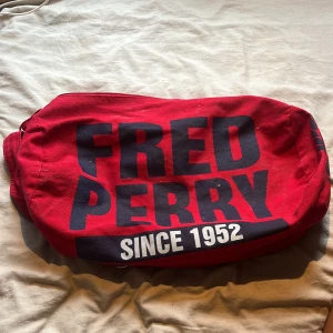 Röd axelväska Fred Perry - Stor röd axelväska från Fred Perry med svart tryck och logga på sidorna. Väskan har breda axelband, dragkedja upptill och rymligt innerfack. Perfekt för att bära med sig mycket saker.