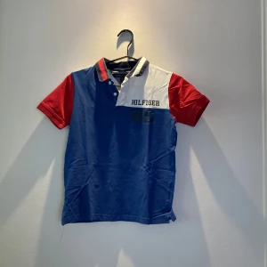 Tommy Hilfiger pikétröja blå/röd/vit - Snygg pikétröja från Tommy Hilfiger i blått, vitt och rött. Klassisk krage med knappar, tryckt 'Hilfiger' och siffran 85 på bröstet. Perfekt för dig som gillar sportig och färgstark stil. Tillverkad i mjuk bomull och har korta ärmar.