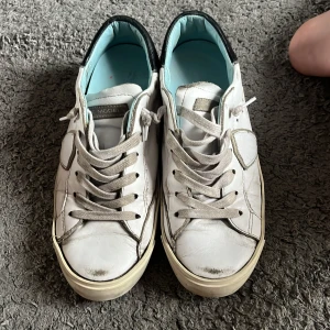 Vita Philippe Model sneakers i skinn - Snygga vita sneakers från Philippe Model i skinn med grå snörning och svarta detaljer vid hälen. Klassisk låg modell med rund tå och platt sula. Insidan är ljusblå och sulan har ett retrovibe. Perfekta för dig som gillar stilrena och trendiga skor.