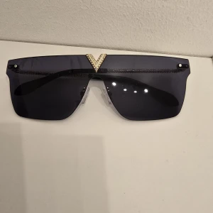Svarta solglasögon med gulddetaljer - Snygga svarta solglasögon med breda, mörka linser och en fet guldfärgad detalj upptill samt ett V-format gulddesign på fronten. Glasögonen har en modern shield-form och näskuddar för extra komfort. Perfekta för dig som vill sticka ut.