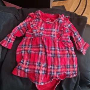 Rutig röd babyklänning från H&M - Supersöt röd rutig babyklänning från H&M i ekologisk bomull. Klänningen har långa ärmar, volangdetaljer vid axlarna och praktiska tryckknappar i grenen. Perfekt för små stunder med extra stil.