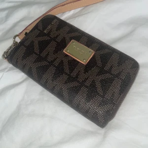 Brun plånbok från Michael Kors - Snygg brun plånbok från Michael Kors med MK-logga. Plånboken har dragkedja runtom, avtagbar handledsrem och flera fack inuti med guldig insida💕Nästan aldrig använt, original pris 850kr. 