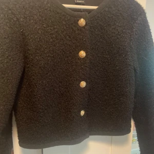 Svart kofta med guldknappar Gina Tricot - Snygg svart kofta från Gina Tricot i storlek S. Koftan är stickad med en fluffig känsla och har fyra dekorativa guldknappar framtill. Perfekt för lager-på-lager och ger en chic vibe till din outfit. Funkar både att ha över en tröja under eller bara som den är. Något tjockar och ”stelare” tyg vilket gör att den känns lyxigare.