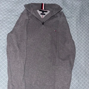 Grå half zip tröja Tommy Hilfiger - Grå långärmad tröja från Tommy Hilfiger med half zip och klassisk liten logga på bröstet. Kragen har röd, vit och marinblå randig detalj. Tillverkad i mjuk premium bomull. Skriv för fler frågor eller bilder i chatten tack!