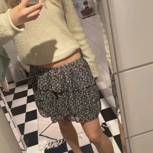 Blommig volangkjol i svart och blått - Supersöt kort kjol med blommigt mönster i svart, blått och vitt. Kjolen har volanger och en luftig känsla, perfekt för varma dagar. Resår i midjan gör den bekväm och enkel att bära. Passar perfekt till en stickad tröja eller en enkel topp.