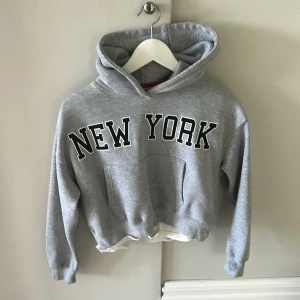 Grå hoodie med New York-tryck från Zara - Grå hoodie från Zara med stor svartvit New York-text på bröstet. Tröjan har huva, magficka och en vit detalj i nederkanten som ger en lager-på-lager-look. Mjuk insida och klassisk passform, perfekt för chill dagar.