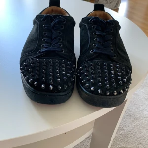 Mörkblå Louboutin sneakers med nitar - Snygga mörkblå sneakers från Christian Louboutin i mocka med coola svarta nitar på tån och klassisk röd sula. Skorna har snörning och rund tå, samt en lyxig känsla med läderfoder. Perfekta för dig som vill sticka ut med en edgy stil. Skriv om ni undrar något