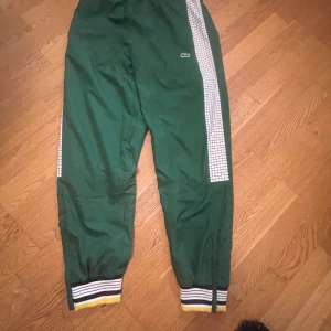 HELT NYA LACOSTE BYXOR - Snygga gröna trackpants från Lacoste med vita rutiga paneler längs sidorna och klassisk krokodillogga. Byxorna har elastiska muddar med svarta, vita och gula ränder nertill. Perfekta för en sportig och retro vibe. Den är helt ny och oanvänd, prislappen borta då min syster råkade ta bort den. Men de är helt nya bortsett från de och inte använda och de är mycket unika och snygga.
