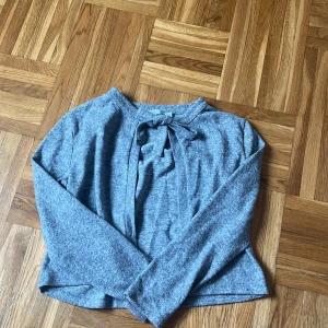 Grå långärmad tröja med knyt - Mysig grå långärmad tröja med knyt framtill i halsen. Tröjan har en enkel och stilren design, perfekt för lager-på-lager och passar till både jeans och kjol. Materialet känns mjukt och bekvämt mot huden.