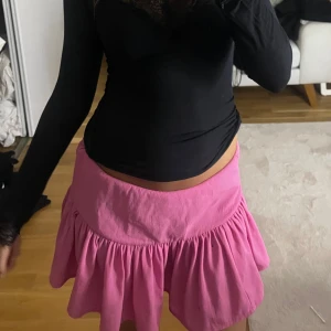 Rosa volangkjol med utsvängd form - Supersöt rosa kortkjol med volang och utsvängd passform. Kjolen har en hög midja och är sydd i ett mjukt bomullstyg som känns skönt mot huden. Perfekt för dig som vill ha en färgglad och lekfull look.