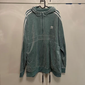 Blå velour hoodie från Adidas XL - Säljer en oversized blå hoodie från Adidas i mjuk velour med klassiska vita ränder på axlarna och Adidas-logga på bröstet. Tröjan har huva, half zip och dragsko nertill. Bekväm och i princip oanvänd