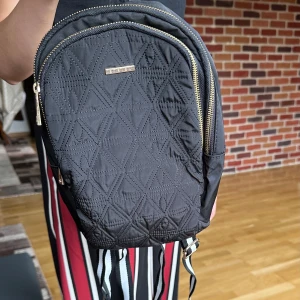 Svart quiltad ryggsäck från LCWaikiki - Snygg svart ryggsäck från LCWaikiki med quiltat geometriskt mönster på framsidan och guldfärgade dragkedjor. Axelremmarna är randiga i svart, vitt och grått. Perfekt storlek för vardagens alla prylar och har en modern look.