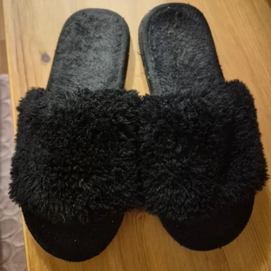 Svarta fluffiga sandaler - Mysiga svarta sandaler med fluffig ovandel och platt sula. Perfekta att glida runt i hemma eller när du vill ha något bekvämt och stilrent. Snyggt helsvart utseende och mjuk känsla tack vare det lurviga materialet.