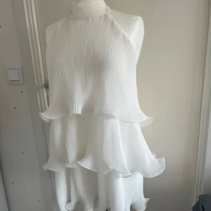 Vit plisserad volangklänning med knyt - Superfin vit kortklänning med plisserat tyg och flera volanger. Hög hals med knyt i nacken och öppen rygg. 