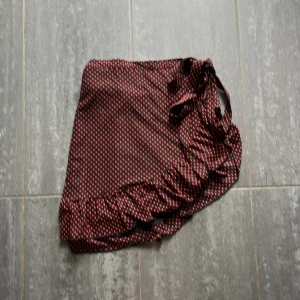 Prickig omlottkjol från Saint Tropez - Supersöt svart kortkjol från Saint Tropez med röda prickar. Kjolen har omlottmodell med knyt i sidan och volang längs nederkanten.