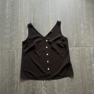 Svart linnetopp med knappar Vero Moda - Svart linnetopp från Vero Moda med v-ringning både fram och bak. Toppen har dekorativa guldiga knappar framtill och är ärmlös med en rak, något lös passform. 