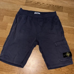 Mörkblå shorts från Stone Island - Snygga mörkblå shorts från Stone Island Junior med klassisk logga på benet. Modellen har en cargoficka på sidan och elastisk midja för skön passform. Perfekta för sommaren och riktigt street. Materialet känns mjukt och är troligtvis bomull.