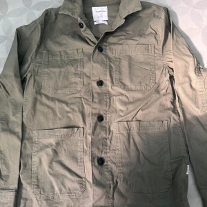 Grön Overshirt från Lindbergh - Stilren beige overshirt från Lindbergh med fyra stora fickor framtill och svarta knappar. Jackan har klassisk krage och är tillverkad i bomull, perfekt för lager-på-lager. Enkel och clean look som passar till det mesta.
