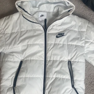 Vit pufferjacka från Nike - Snygg vit pufferjacka från Nike med svart logga på bröstet och svarta dragkedjor. Jackan har huva och två fickor med dragkedja. Perfekt för kyliga dagar när du vill hålla stilen och värmen. Passar både M/L