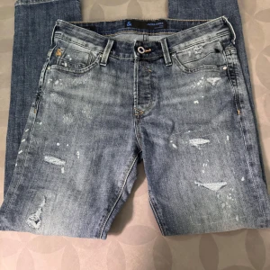 Jack & Jones slitna blå jeans 31/32 - Snygga blå jeans från Jack & Jones i storlek 31/32. Jeansen har en tvättad look med slitningar, lagade detaljer och coola färgstänk. Klassisk femficksmodell med gylf och bälteshällor. Perfekta för en avslappnad streetstil.