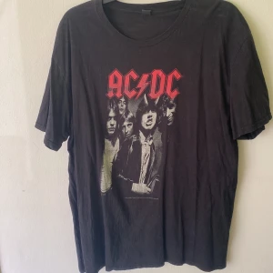 Svart AC/DC t-shirt med tryck - Svart t-shirt med stort AC/DC-tryck i rött och grått på bröstet. Framsidan har ett ikoniskt bandmotiv med medlemmarna i svartvitt. Klassisk rund hals och korta ärmar. Perfekt för dig som gillar rock och vill sticka ut med en cool bandtröja.