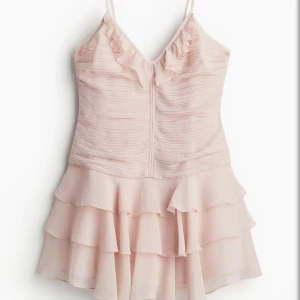 Ljusrosa volangklänning med smala axelband - Supersöt ljusrosa kortklänning med smala axelband, v-ringning och volanger både upptill och nertill. Klänningen har flera lager volang i kjolen och en lätt rynkad struktur i tyget. Perfekt för dig som gillar romantisk och feminin stil.
