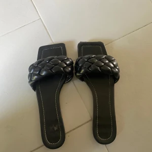 Svarta flätade sandaler med fyrkantig tå - Snygga svarta sandaler med bred, flätad rem över foten och fyrkantig tå. Skorna har platt sula och vita kontrastsömmar längs kanterna. Perfekta för en trendig och avslappnad look under varma dagar.