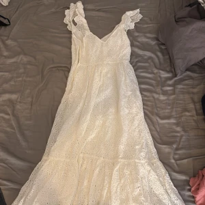Vit långklänning med spetsdetaljer - Romantisk vit långklänning med broderade spetsdetaljer och volangkant. Klänningen har v-ringning och breda axelband med volang. Perfekt för sommarens alla tillfällen och ger en riktigt drömmig känsla.