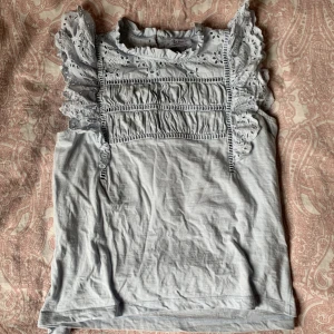 Ljusblå blus med volang från Lindex - Supersöt ljusblå blus från Holly & Whyte by Lindex. Blusen har broderade spetsdetaljer, volang vid axlarna och halsen samt en liten guldig knapp i nacken. Knappt använd, mycket bra skick!