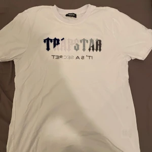 Vit t-shirt från Trapstar - Säljer en vit t-shirt från Trapstar med tryck i svart och grått på framsidan. T-shirten har en klassisk rund hals och korta ärmar. Perfekt för en avslappnad stil.