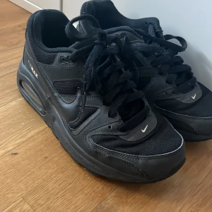 Svarta Nike Air Max sneakers - Säljer ett par svarta Nike Air Max sneakers med klassisk design och synlig Air Max-dämpning i sulan. Skorna har snörning och är tillverkade i syntetmaterial med Nike-logga på sidan och hälen.