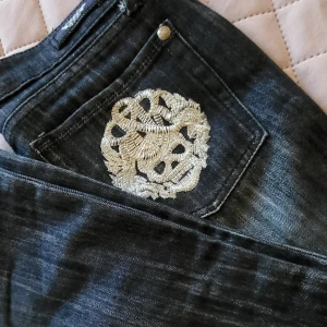 Svarta jeans från Rock & Republic - Snygga svarta jeans från Rock & Republic med coolt broderi på bakfickan. Jeansen har en klassisk femficksdesign och en knappgylf. Perfekta för en edgy look!