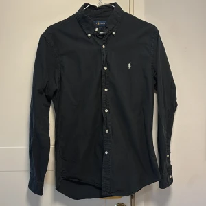 Svart skjorta från Ralph Lauren - Snygg svart skjorta från Ralph Lauren i slim fit. Skjortan har långa ärmar och klassiska knappar framtill. Den ikoniska loggan är broderad på bröstet. Perfekt för en stilren look.