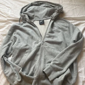 Grå hoodie från Nike - Säljer en grå hoodie från Nike med dragkedja och huva. Tröjan har långa ärmar och en liten Nike-logga på bröstet. 
