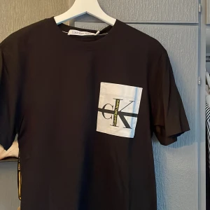 Svart t-shirt från Calvin Klein Jeans - Snygg svart t-shirt från Calvin Klein Jeans med en vit bröstficka som har en stor logotyp i svart och gult. Perfekt för en stilren och modern look.