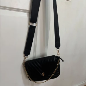 Svart quiltad axelväska med kedja - Snygg svart axelväska i quiltat mönster med guldfärgad kedjedetalj. Väskan har en justerbar axelrem och ett elegant spänne framtill. Perfekt för att ge en stilren touch till din outfit.