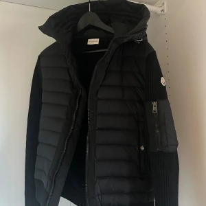 Svart cardigan jacka från Moncler - Säljer en snygg svart cardigan jacka från Moncler med stickade ärmar och en quiltad kropp. Jackan har en dragkedja framtill och en huva för extra stil och värme. Perfekt för kyligare dagar!