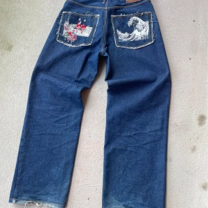 Blå jeans med broderade fickor - Snygga blå jeans med unika broderade detaljer på bakfickorna. Ena fickan har ett vågmotiv och den andra ett färgglatt mönster. Jeansen har en rak passform och är perfekta för en avslappnad stil.