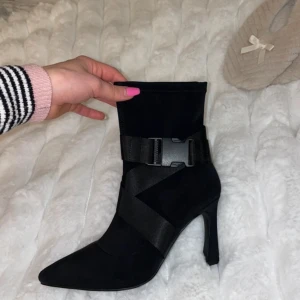 Svarta stövletter från NA-KDxHannaLicious - Snygga svarta stövletter från NA-KD med spetsig tå och klack. Skorna har en unik design med breda band och spänne runt ankeln. Perfekta för att ge en stilren touch till din outfit.