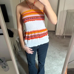Färgglad axelbandslös topp - Säljer en färgglad axelbandslös topp med halterneck-band. Toppen har ett mönster i rött, gult och orange på en vit bakgrund. Perfekt för sommarens varma dagar!