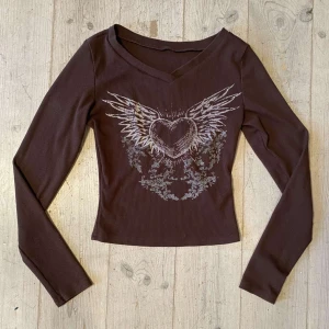 Brun ribbad långärmad topp med tryck - Snygg brun ribbad topp med ett coolt tryck av ett hjärta med vingar på framsidan. Toppen har en v-ringad hals och långa ärmar, perfekt för en avslappnad stil.