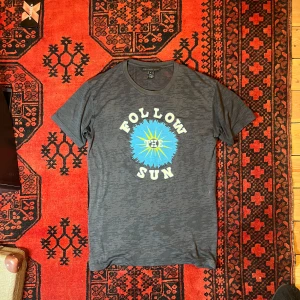 Grå t-shirt från Marc by Marc Jacobs - Säljer en grå t-shirt från Marc by Marc Jacobs med texten 'Follow the Sun' i vitt och en blå sol i mitten. Perfekt för en avslappnad stil.