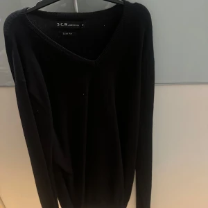 Svart tröja från S.C.W. - Säljer en stilren svart tröja från S.C.W. i slim fit-modell. Tröjan har en v-ringad design och långa ärmar, perfekt för en elegant och avslappnad look.