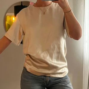 Beige satintopp med dragkedja - Säljer en stilren beige t-shirt i ett tjockare satinmaterial med korta ärmar och en dragkedja i ryggen. Passar supersnyggt med en jeanskjol nu till våren! Den har en liten fläck vid urringningen, den borde dock enkelt gå bort efter en tvätt🤍