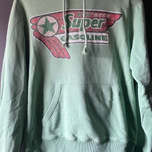 Grön hoodie med tryck - Säljer en grön hoodie med ett stort tryck på framsidan. Hoodien har en känguruficka och justerbar dragsko i huvan. Perfekt för en avslappnad stil.