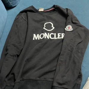 Svart sweatshirt från Moncler - Snygg svart sweatshirt från Moncler med deras ikoniska logga broderad på bröstet och en patch på ärmen. Tröjan har en rund halsringning och ribbade muddar vid ärmslut och nederkant. Perfekt för en stilren look.