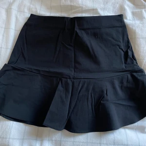 Svart volangkjol från Zara - Snygg svart kortkjol från Zara med volangdetalj. Perfekt för att ge en stilren look. Har inbyggda shorts och en dragkedja. Säljer endast då den inte kommer till användning längre. Storlek S nypris 349 kr.