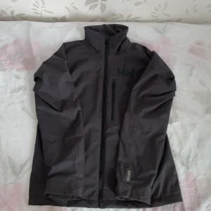 Svart vindjacka från Helly Hansen - Säljer en stilren svart vindjacka från Helly Hansen. Jackan har en hög krage och dragkedja framtill samt en praktisk bröstficka med dragkedja. Perfekt för blåsiga dagar.