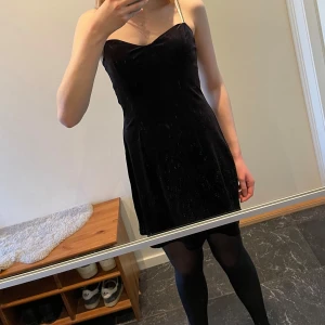 Svart klänning med strassprydda axelband från H&M - Snygg svart klänning från H&M med strassprydda axelband. Klänningen har en tight passform upptill men en står ut lite från midjan, den är kort men går ändå ner en bra bit över vaderna. Tyget är sammetsliknande. Perfekt för en kväll ute eller en fest!